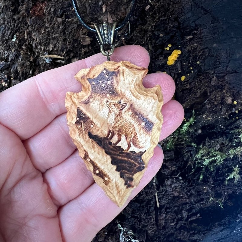 wood-carved arrowhead pendant featuring a fox looking at the aurora borealis